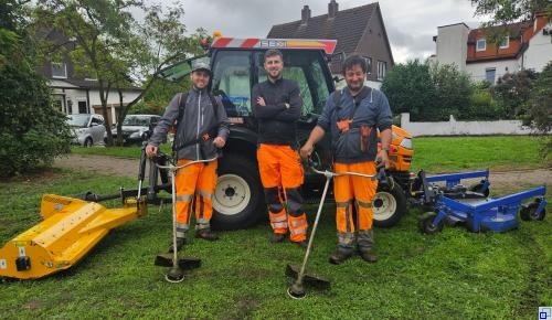 Drei Männer mit orangenen Arbeitshosen stehen vor einem Traktor mit angehängtem Mähwerk. Zwei von Ihnen haben Freischneider in den Händen. 