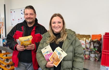 Links steht eine Frau mit zwei Packungen Leckerlis in der Hand, rechts daneben ein Mann, ebenfalls mit zwei Futterpackungen in der Hand. Beide lächeln in die Kamera.